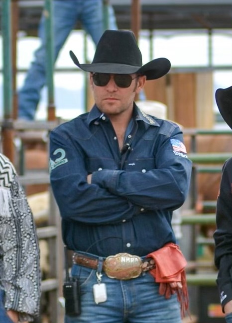 Man in blue shirt, black cowboy hat and sunglasses standing with arms crossed